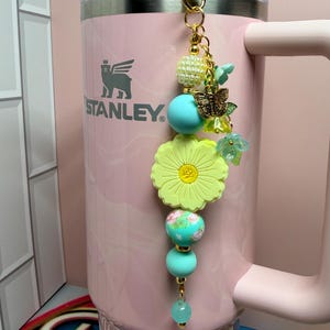 May include: A light pink Stanley tumbler with a decorative beaded charm attached to the handle. The charm features a large yellow flower bead, turquoise silicone spheres, a gold butterfly charm, and small green floral accents on a gold chain.
