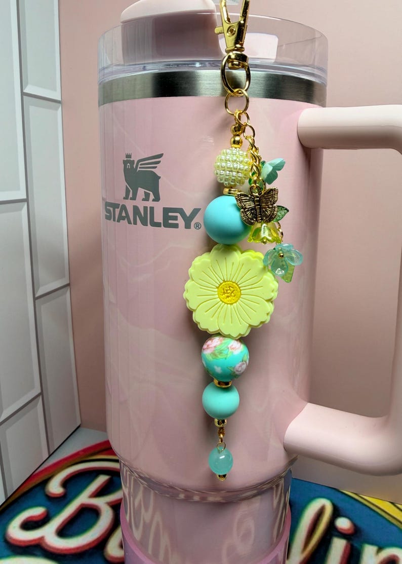 May include: A light pink Stanley tumbler with a decorative beaded charm attached to the handle. The charm features a large yellow flower bead, turquoise silicone spheres, a gold butterfly charm, and small green floral accents on a gold chain.