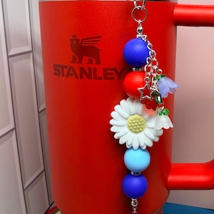 Op de afbeelding: Een rode Stanley-beker met een blauwe rietje en een decoratieve bedel. De bedel heeft blauwe, rode en witte kralen, een witte madelief en kleine bloemaccenten. De beker heeft het Stanley-logo.
