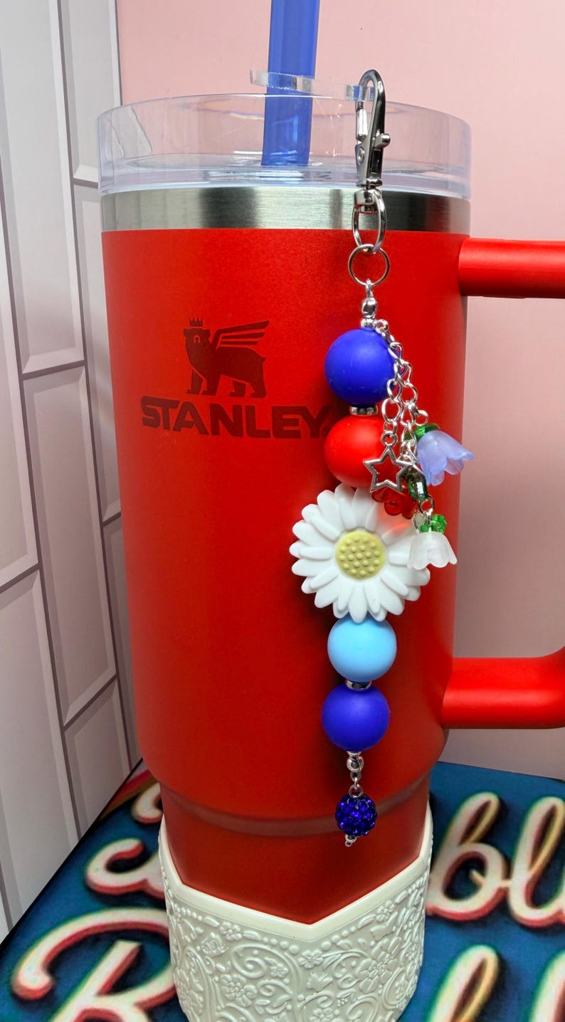 Op de afbeelding: Een rode Stanley-beker met een blauwe rietje en een decoratieve bedel. De bedel heeft blauwe, rode en witte kralen, een witte madelief en kleine bloemaccenten. De beker heeft het Stanley-logo.