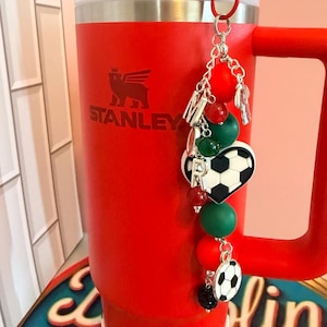 Puede incluir: Un vaso Stanley de color rojo brillante con tapa y asa blancas, que presenta un accesorio decorativo colgante unido al asa. El adorno incluye cuentas de letras plateadas, cuentas de silicona rojas y verdes, y dos dijes de balones de f&uacute;tbol.