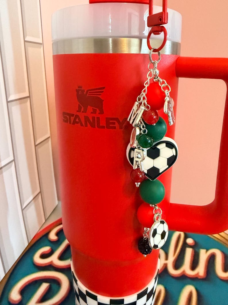 Puede incluir: Un vaso Stanley de color rojo brillante con tapa y asa blancas, que presenta un accesorio decorativo colgante unido al asa. El adorno incluye cuentas de letras plateadas, cuentas de silicona rojas y verdes, y dos dijes de balones de f&uacute;tbol.