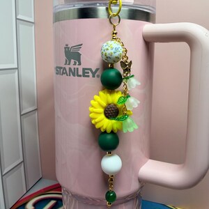 May include: A light pink Stanley tumbler with a handle and a decorative keychain attached to the lid. The keychain features a yellow sunflower charm, a white bead with yellow floral patterns, dark green and white beads, and small bell-shaped charms.