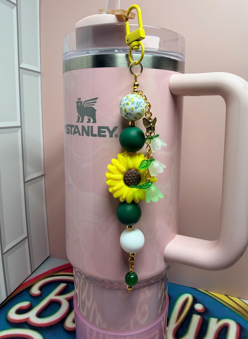 May include: A light pink Stanley tumbler with a handle and a decorative keychain attached to the lid. The keychain features a yellow sunflower charm, a white bead with yellow floral patterns, dark green and white beads, and small bell-shaped charms.