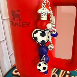 Puede incluir: Un vaso rojo brillante con asa presenta un dije decorativo sujeto en la parte superior. El dije incluye un colgante de camiseta de f&uacute;tbol plateado, un bal&oacute;n de f&uacute;tbol en forma de coraz&oacute;n, un bal&oacute;n redondo y letras plateadas que forman LISA.