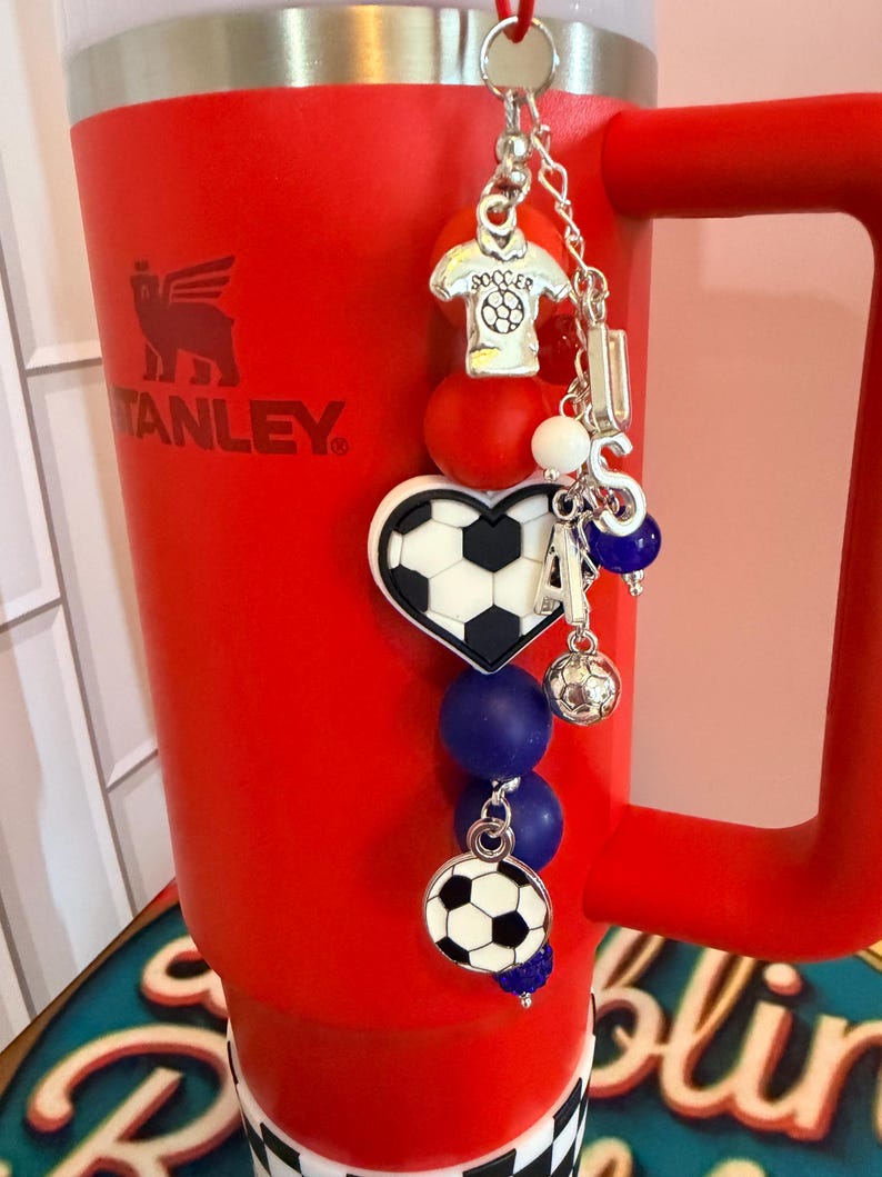 Puede incluir: Un vaso rojo brillante con asa presenta un dije decorativo sujeto en la parte superior. El dije incluye un colgante de camiseta de f&uacute;tbol plateado, un bal&oacute;n de f&uacute;tbol en forma de coraz&oacute;n, un bal&oacute;n redondo y letras plateadas que forman LISA.