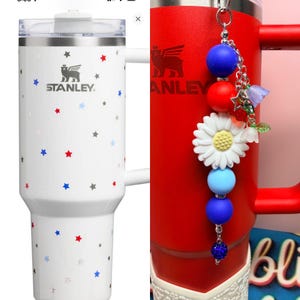 Op de afbeelding: Witte Stanley-beker met een handvat, versierd met rode, blauwe en grijze sterren. Het Stanley-logo is zichtbaar. Een rode Stanley-mok met een decoratieve bedel met blauwe, rode en witte kralen, een madeliefje en een ster.
