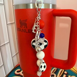 Puede incluir: Un vaso Stanley rojo con asa y tapa blanca, que presenta un accesorio colgante. El dije incluye cuentas de letras plateadas que forman ENG, cuentas de silicona redondas en naranja, azul y blanco, y dijes con tem&aacute;tica de bal&oacute;n de f&uacute;tbol.