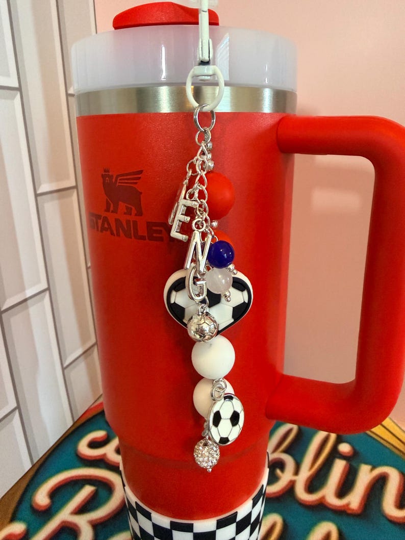 Puede incluir: Un vaso Stanley rojo con asa y tapa blanca, que presenta un accesorio colgante. El dije incluye cuentas de letras plateadas que forman ENG, cuentas de silicona redondas en naranja, azul y blanco, y dijes con tem&aacute;tica de bal&oacute;n de f&uacute;tbol.