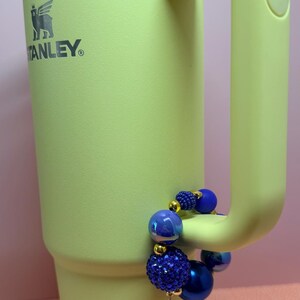 Op de afbeelding: Een lichtgroene Stanley-beker met een handvat en een doorzichtig deksel. De beker heeft het Stanley-logo. Een blauwe kralenarmband met een gouden vliegtuigbedel is aan het handvat bevestigd.