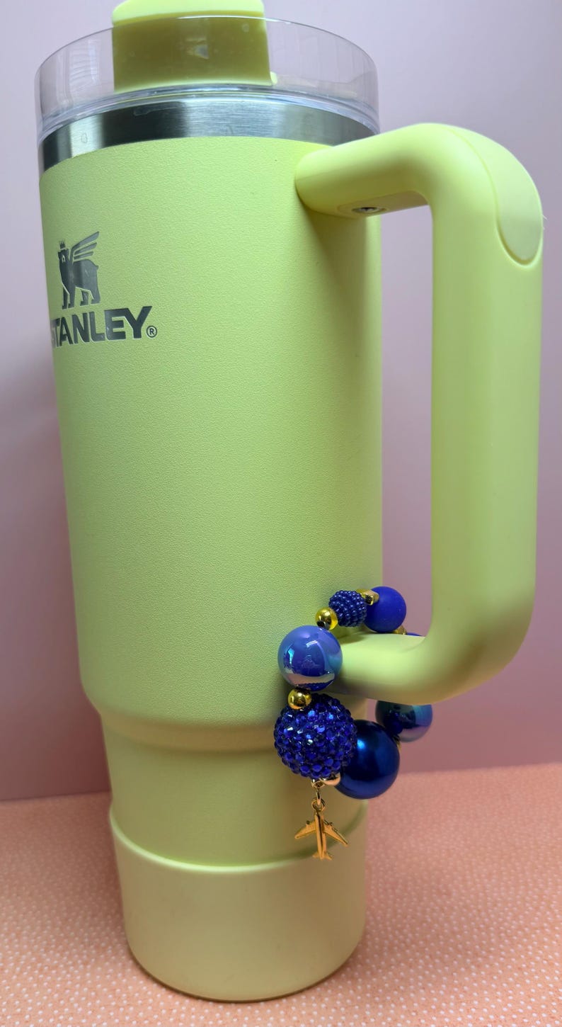 Op de afbeelding: Een lichtgroene Stanley-beker met een handvat en een doorzichtig deksel. De beker heeft het Stanley-logo. Een blauwe kralenarmband met een gouden vliegtuigbedel is aan het handvat bevestigd.