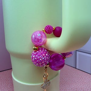 Op de afbeelding: Een roze kralenarmband met een gouden ballerina-bedel. De armband heeft grote, ronde kralen in roze en fuchsia tinten, met kleinere gouden accentkralen. De ballerina-bedel is goudkleurig met een strass-tutu.
