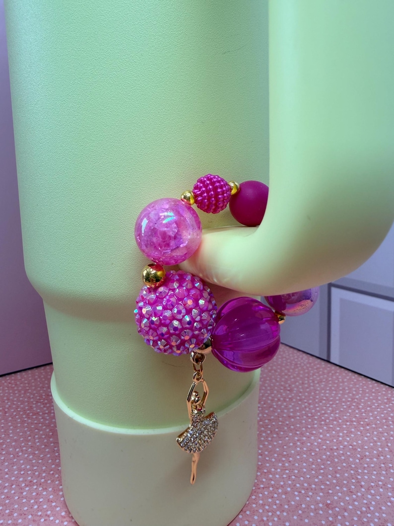 Op de afbeelding: Een roze kralenarmband met een gouden ballerina-bedel. De armband heeft grote, ronde kralen in roze en fuchsia tinten, met kleinere gouden accentkralen. De ballerina-bedel is goudkleurig met een strass-tutu.