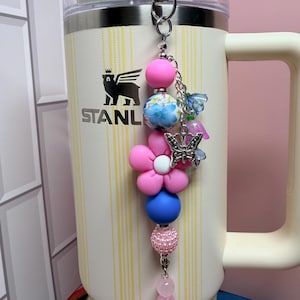 May include: A cream-colored Stanley tumbler with vertical yellow stripes and a black bear logo, featuring a decorative beaded keychain. The keychain includes pink, blue, and floral beads, a pink flower-shaped bead, a silver butterfly charm, and small blue and pink flower charms attached to a silver metal clasp.