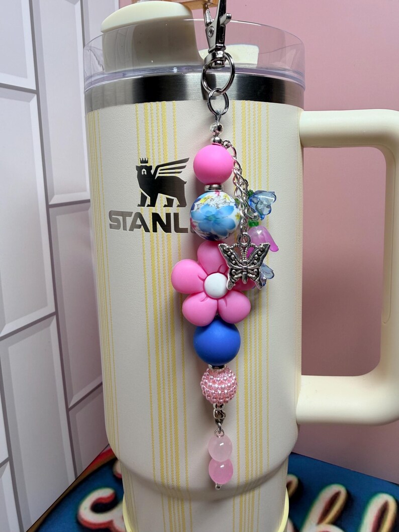 May include: A cream-colored Stanley tumbler with vertical yellow stripes and a black bear logo, featuring a decorative beaded keychain. The keychain includes pink, blue, and floral beads, a pink flower-shaped bead, a silver butterfly charm, and small blue and pink flower charms attached to a silver metal clasp.