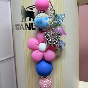 May include: A decorative tumbler charm featuring a vertical arrangement of beads including a solid pink sphere, a blue and white floral patterned bead, a pink flower-shaped bead, a solid blue sphere, and a textured pink bead with a silver butterfly charm.