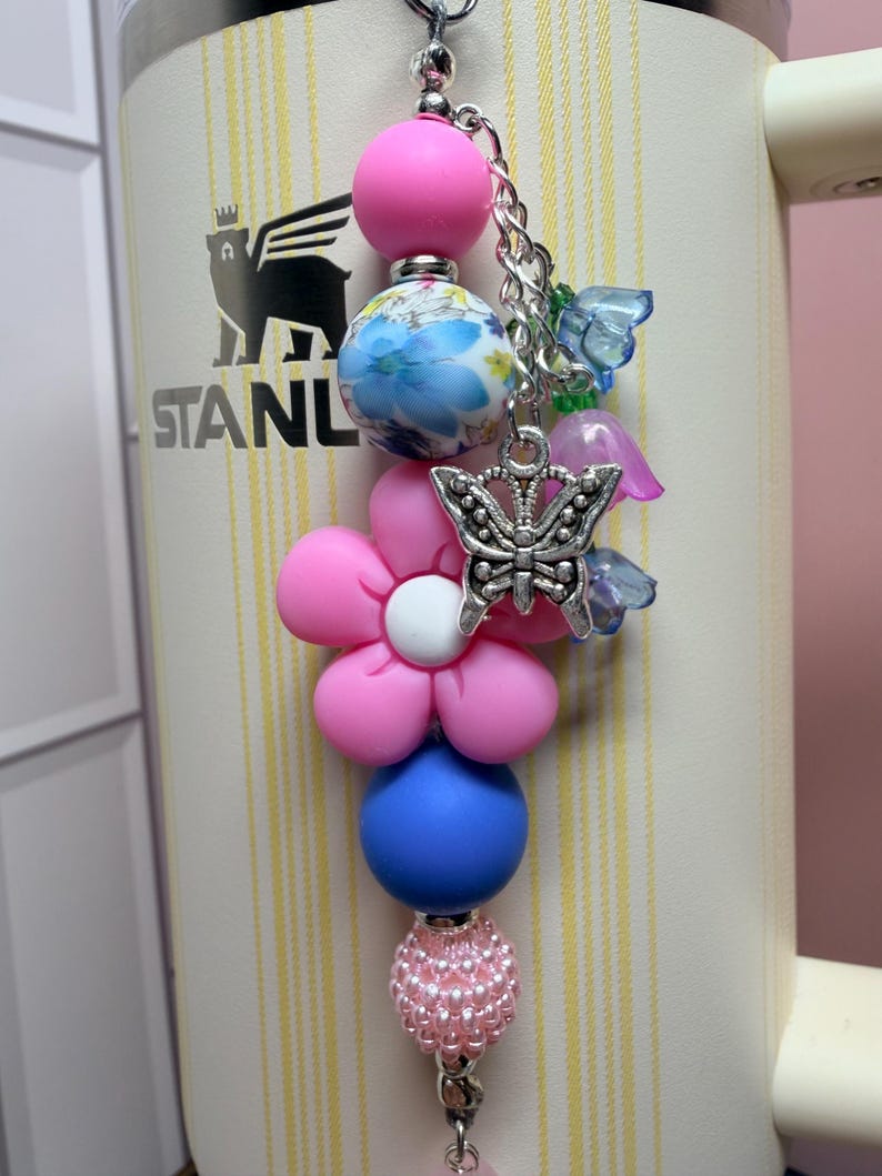 May include: A decorative tumbler charm featuring a vertical arrangement of beads including a solid pink sphere, a blue and white floral patterned bead, a pink flower-shaped bead, a solid blue sphere, and a textured pink bead with a silver butterfly charm.