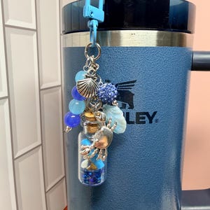 May include: A blue lobster clasp keychain featuring a small glass bottle filled with blue glitter and beads. The charm includes silver-toned metal accents shaped like a crab and a seashell, along with blue beads and a textured blue sphere.