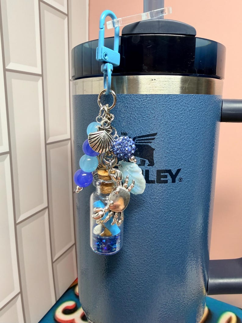 May include: A blue lobster clasp keychain featuring a small glass bottle filled with blue glitter and beads. The charm includes silver-toned metal accents shaped like a crab and a seashell, along with blue beads and a textured blue sphere.