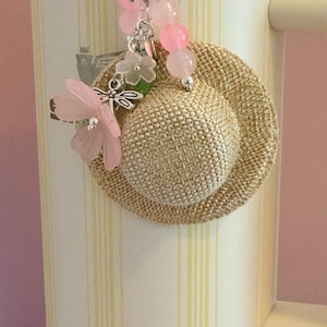May include: A cream travel mug with yellow stripes, a small woven hat charm, and a beaded charm with pink beads, a dragonfly, and a pink flower.