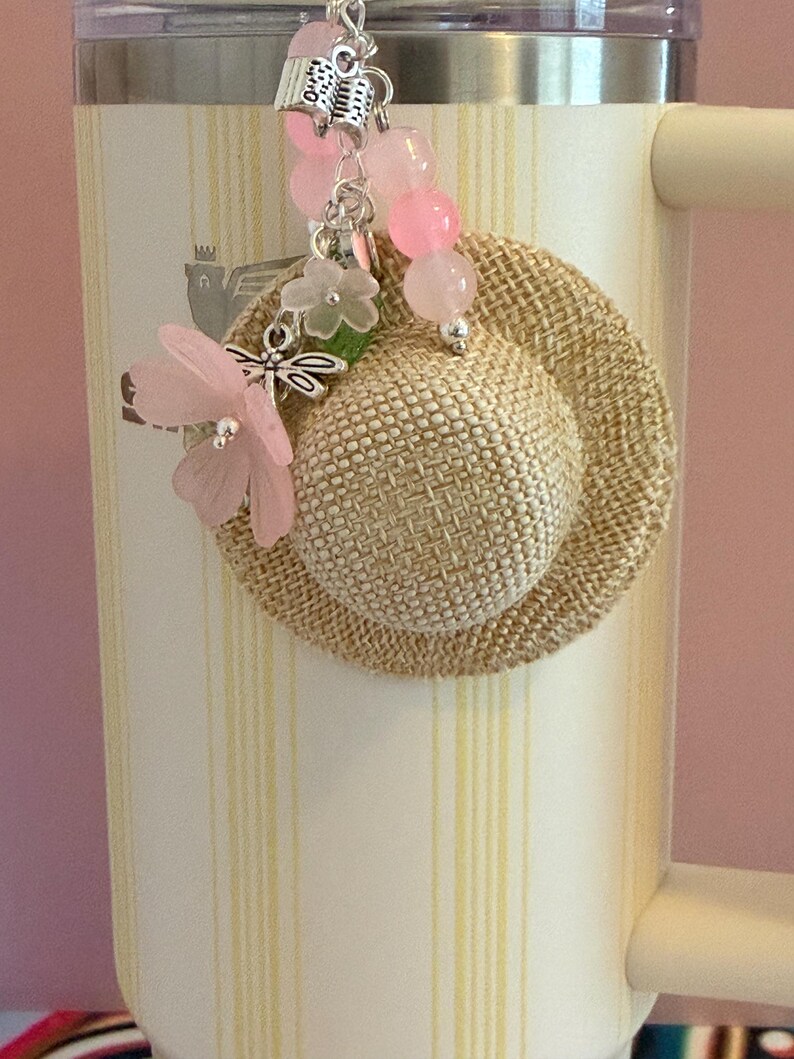 May include: A cream travel mug with yellow stripes, a small woven hat charm, and a beaded charm with pink beads, a dragonfly, and a pink flower.