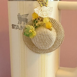May include: A decorative charm with a small woven hat, yellow beads, and green floral details. A gold butterfly and lace element are suspended above the hat. The charm is attached to a cream-coloured tumbler.