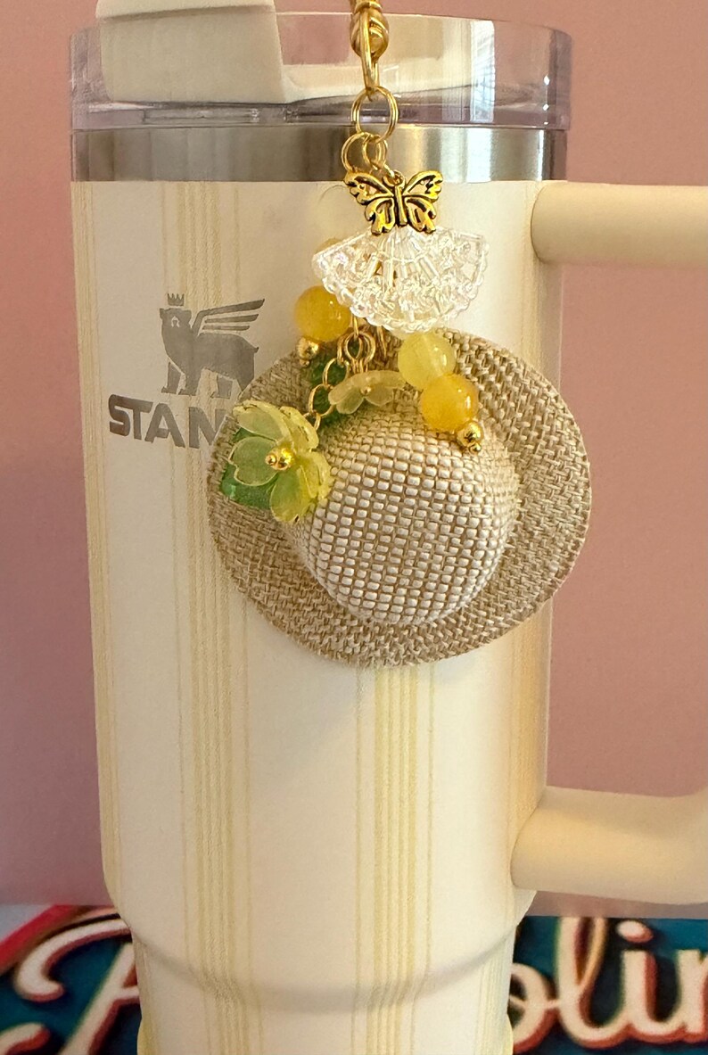 May include: A decorative charm with a small woven hat, yellow beads, and green floral details. A gold butterfly and lace element are suspended above the hat. The charm is attached to a cream-coloured tumbler.