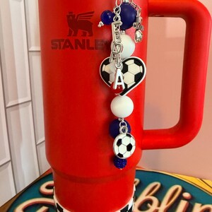 Puede incluir: Un vaso Stanley de color rojo brillante con una base de cuadros blancos y negros y un dije decorativo de cuentas. El dije incluye cuentas azules y blancas, un dije de bal&oacute;n de f&uacute;tbol en forma de coraz&oacute;n, un bal&oacute;n redondo y letras plateadas.