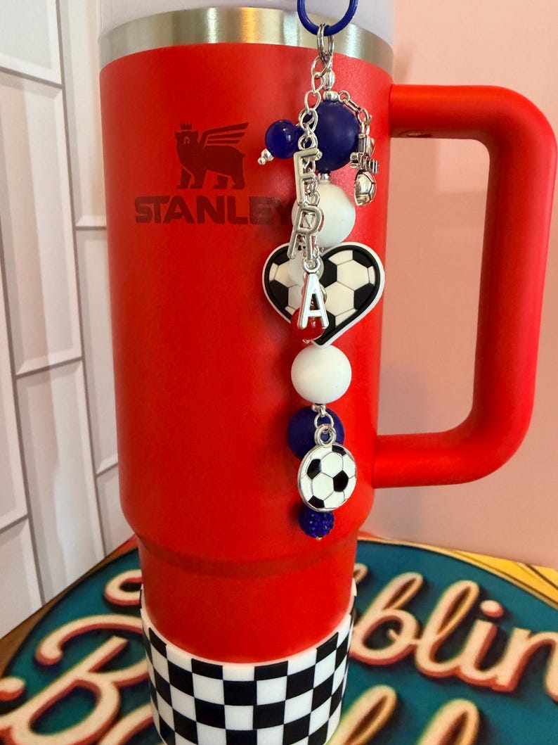 Puede incluir: Un vaso Stanley de color rojo brillante con una base de cuadros blancos y negros y un dije decorativo de cuentas. El dije incluye cuentas azules y blancas, un dije de bal&oacute;n de f&uacute;tbol en forma de coraz&oacute;n, un bal&oacute;n redondo y letras plateadas.