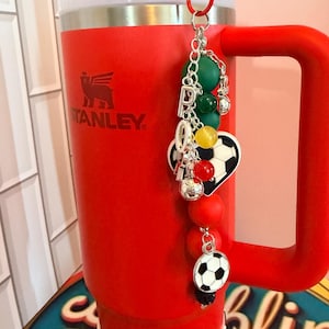 Puede incluir: Un vaso Stanley de color rojo brillante con asa y tapa blanca, que presenta un llavero decorativo sujeto en la parte superior. El llavero incluye dijes de letras plateadas que forman la palabra PRO, cuentas redondas verdes, amarillas y rojas, y balones.