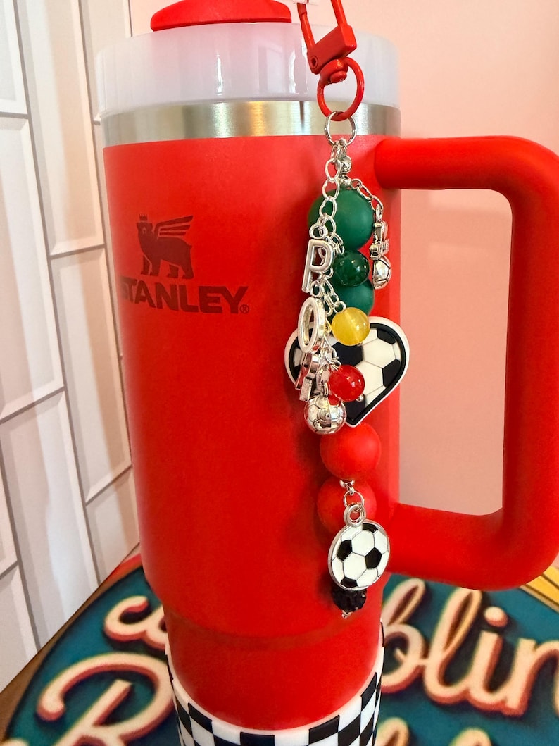 Puede incluir: Un vaso Stanley de color rojo brillante con asa y tapa blanca, que presenta un llavero decorativo sujeto en la parte superior. El llavero incluye dijes de letras plateadas que forman la palabra PRO, cuentas redondas verdes, amarillas y rojas, y balones.