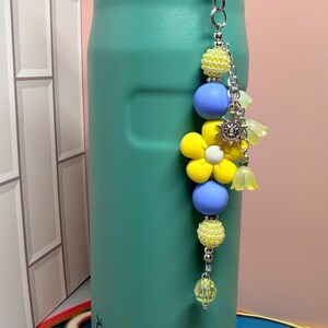 May include: A teal Stanley water bottle with a decorative beaded charm attached to the handle. The charm features yellow textured beads, smooth periwinkle blue spheres, a large yellow flower bead, a silver sun charm, and small translucent yellow flower-shaped beads.