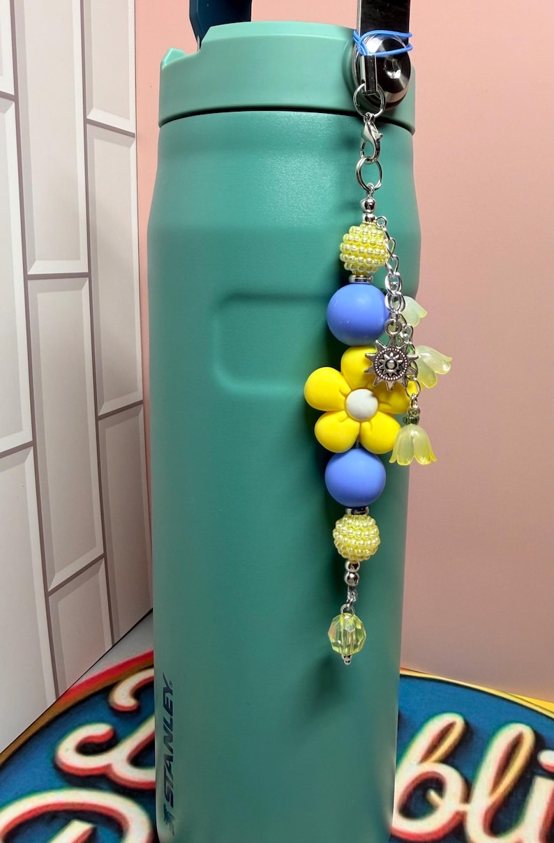 May include: A teal Stanley water bottle with a decorative beaded charm attached to the handle. The charm features yellow textured beads, smooth periwinkle blue spheres, a large yellow flower bead, a silver sun charm, and small translucent yellow flower-shaped beads.
