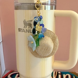 May include: A decorative keychain charm with a miniature straw hat, sunglasses, and a gold butterfly. The charm is decorated with blue and green floral accents and hangs from a gold clasp. The charm is attached to a cream-coloured Stanley tumbler.