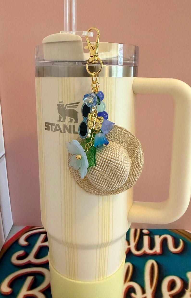 May include: A decorative keychain charm with a miniature straw hat, sunglasses, and a gold butterfly. The charm is decorated with blue and green floral accents and hangs from a gold clasp. The charm is attached to a cream-coloured Stanley tumbler.