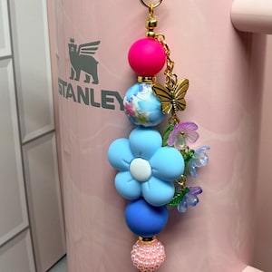 May include: A decorative tumbler charm featuring a gold-tone metal clasp, a bright pink bead, a blue floral patterned bead, a large light blue flower-shaped bead, a matte blue bead, a textured light pink bead, and a gold-tone butterfly charm.