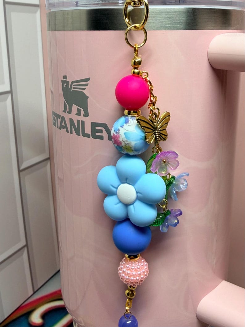 May include: A decorative tumbler charm featuring a gold-tone metal clasp, a bright pink bead, a blue floral patterned bead, a large light blue flower-shaped bead, a matte blue bead, a textured light pink bead, and a gold-tone butterfly charm.
