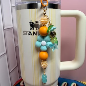 May include: A decorative beaded charm hanging from a cream-colored Stanley tumbler. The charm features orange and pearl-textured beads, a large light blue flower bead, a small gold heart charm, a green leaf charm, and a translucent blue butterfly.