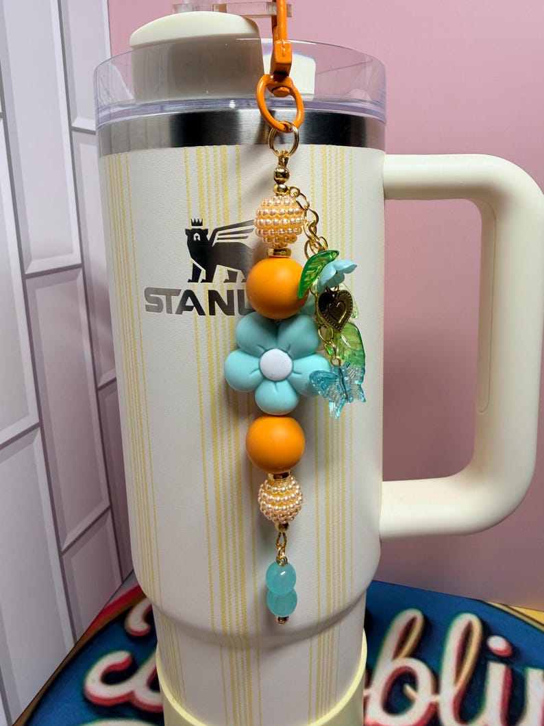 May include: A decorative beaded charm hanging from a cream-colored Stanley tumbler. The charm features orange and pearl-textured beads, a large light blue flower bead, a small gold heart charm, a green leaf charm, and a translucent blue butterfly.