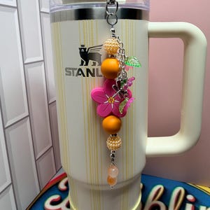 May include: A cream-colored Stanley tumbler with vertical yellow stripes features a decorative beaded charm attached to the lid. The charm includes orange and pearl-textured beads, a bright pink flower-shaped silicone bead, and small green leaf accents.