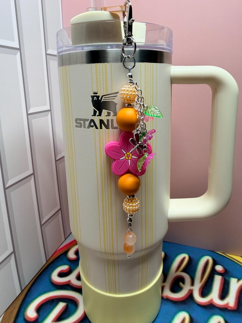 May include: A cream-colored Stanley tumbler with vertical yellow stripes features a decorative beaded charm attached to the lid. The charm includes orange and pearl-textured beads, a bright pink flower-shaped silicone bead, and small green leaf accents.