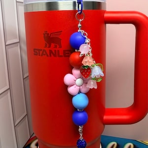 Op de afbeelding: Een rode Stanley-beker met een blauwe rietje en een decoratieve bedel. De bedel heeft blauwe, rode en roze kralen, evenals bloem- en aardbei-accenten. De beker heeft het "STANLEY"-logo.