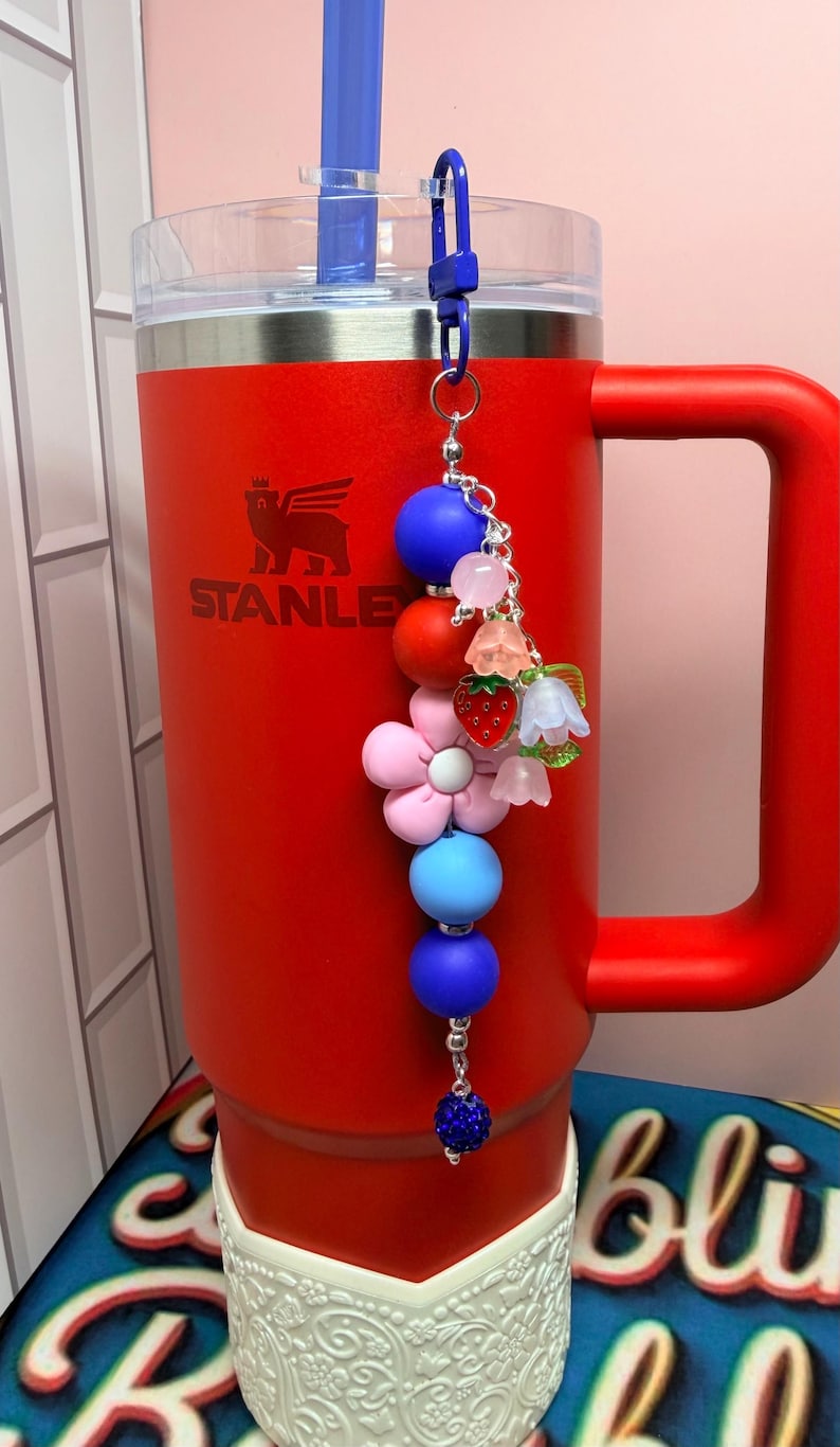 Op de afbeelding: Een rode Stanley-beker met een blauwe rietje en een decoratieve bedel. De bedel heeft blauwe, rode en roze kralen, evenals bloem- en aardbei-accenten. De beker heeft het "STANLEY"-logo.