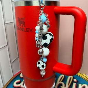 Puede incluir: Un vaso Stanley rojo brillante con asa y base a cuadros blancos y negros. Del tap&oacute;n cuelga un adorno decorativo con cuentas azul claro y blancas, letras plateadas que forman ARG, un bal&oacute;n de f&uacute;tbol en forma de coraz&oacute;n y otros dijes.