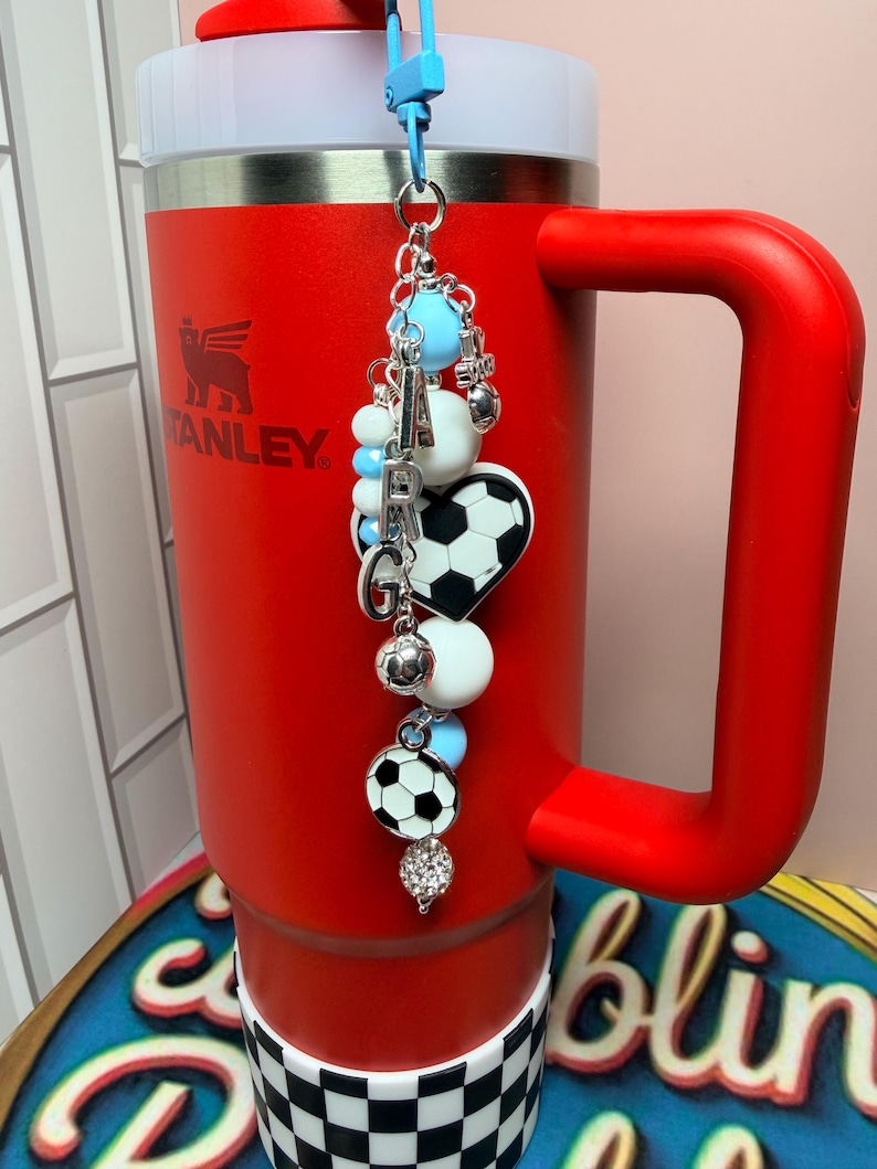 Puede incluir: Un vaso Stanley rojo brillante con asa y base a cuadros blancos y negros. Del tap&oacute;n cuelga un adorno decorativo con cuentas azul claro y blancas, letras plateadas que forman ARG, un bal&oacute;n de f&uacute;tbol en forma de coraz&oacute;n y otros dijes.