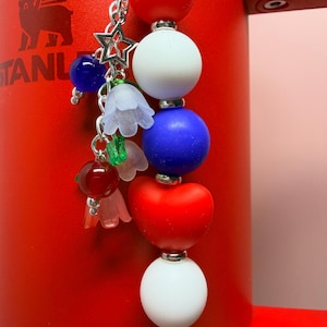 Op de afbeelding: Een patriottische bedel met rode, witte en blauwe kralen, een sterbedel en bloemaccenten. De bedel hangt aan een zilveren ketting tegen een rode achtergrond. Een decoratief accessoire.