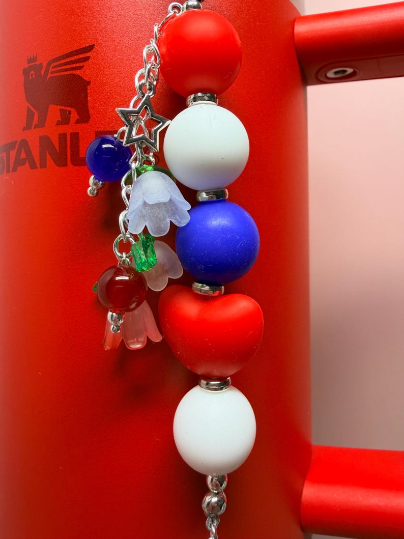 Op de afbeelding: Een patriottische bedel met rode, witte en blauwe kralen, een sterbedel en bloemaccenten. De bedel hangt aan een zilveren ketting tegen een rode achtergrond. Een decoratief accessoire.