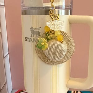 May include: A cream-coloured tumbler with vertical yellow stripes, featuring a decorative charm. The charm includes a miniature straw hat, yellow beads, and a gold butterfly. The tumbler has a handle and the word "STANLEY" is visible.