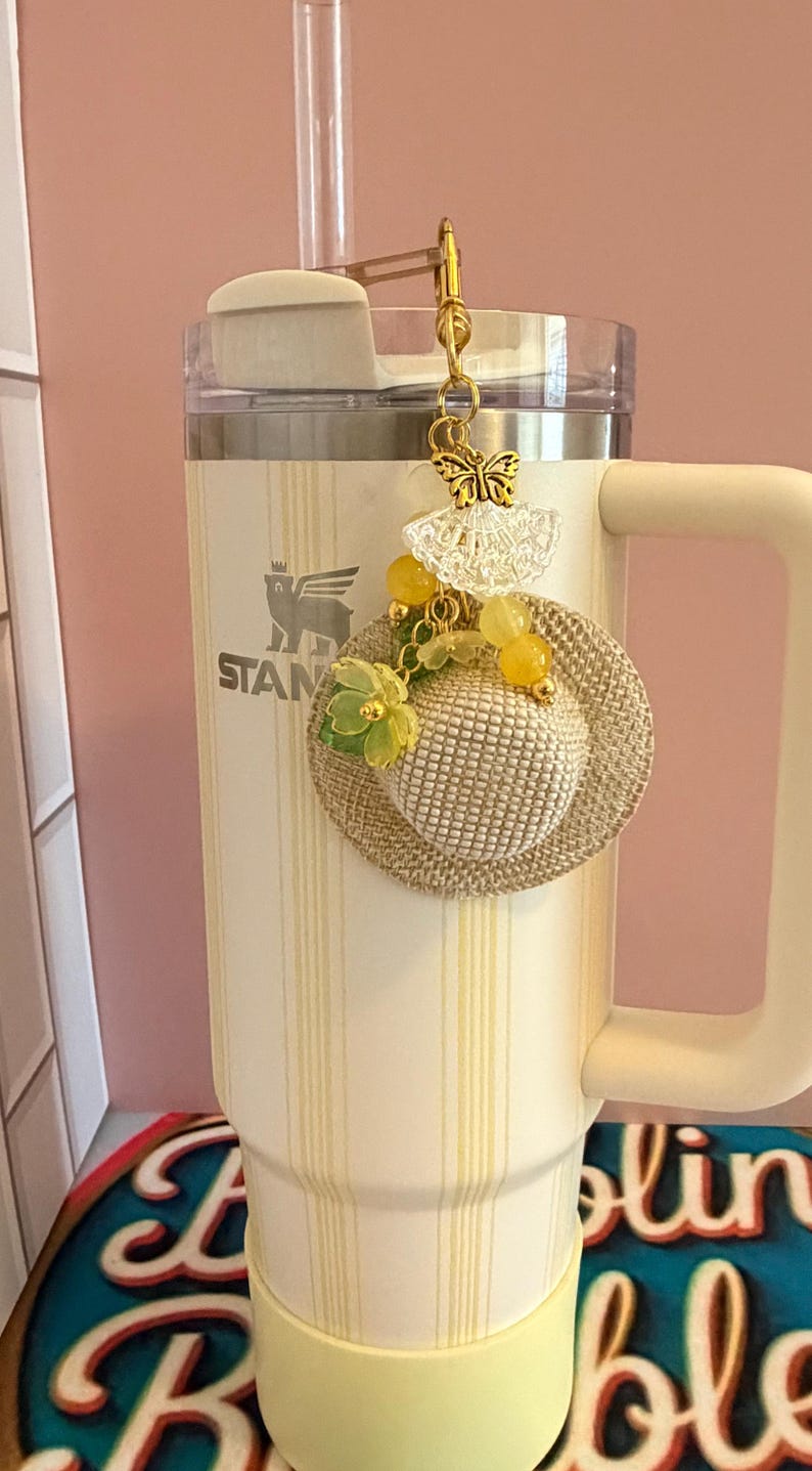 May include: A cream-coloured tumbler with vertical yellow stripes, featuring a decorative charm. The charm includes a miniature straw hat, yellow beads, and a gold butterfly. The tumbler has a handle and the word "STANLEY" is visible.