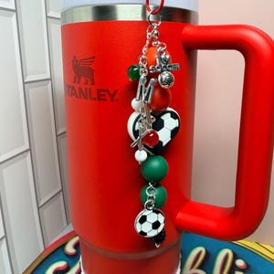 Puede incluir: Un vaso Stanley de color rojo brillante con asa y tapa blanca, que presenta un dije decorativo sujeto en la parte superior. El dije incluye una letra M plateada, un dije de bal&oacute;n de f&uacute;tbol, una cuenta en forma de coraz&oacute;n y cuentas verdes, rojas y blancas.